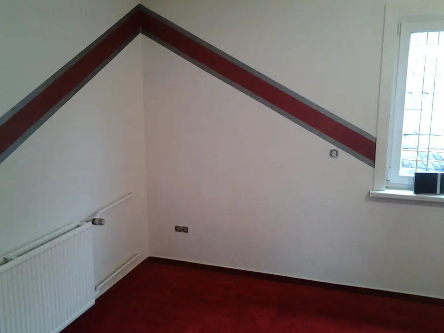 Weiß gestrichenes Dachgeschosszimmer mit umlaufendem rot-grauem Streifenband entlang der Dachschräge